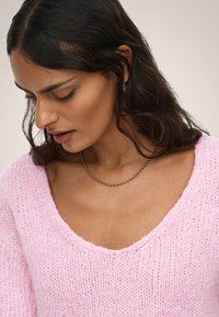 Femme regardant vers le bas, portant un pull rose en maille à col en V, un collier à chaîne dorée et une petite boucle d'oreille anneau dorée sur un fond neutre.