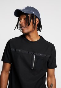 Chemise noire à manches courtes avec un panneau texturé, poche zippée et accent de logo. Associée à une casquette de baseball bleu marine ornée de broderies.