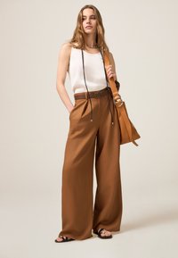 Pantalones marrones de pierna ancha hechos de una tela suave, combinados con una blusa sin mangas blanca y un cinturón marrón. Presenta un bolso de hombro en tono marrón y sandalias negras.