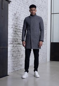 Abrigo gris de cuello alto con silueta recta, llevado sobre pantalones negros y zapatillas blancas, contra una pared de ladrillo blanco.