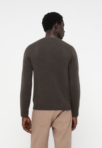 Brun stickad tröja med långa ärmar, rund halsringning och ribbade ärmslut, sedd bakifrån, tillsammans med beige strukturerade byxor.