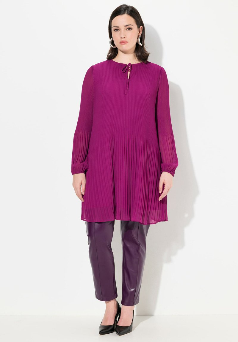 Paarse geplisseerde jurk met lange mouwen en een striknek, gecombineerd met slim-fit paarse leren broek en zwarte spitse hakken. Gladde textuur, modern ontwerp.