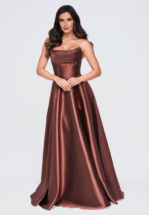 Mujer con cabello largo y oscuro, vistiendo un vestido marrón de satén sin tirantes y largo hasta el suelo, con detalles de lentejuelas en el escote, de pie con las manos entrelazadas.