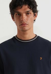 Granatowy sweter z kontrastującym prążkowanym kołnierzem w paski, wykonany z gładkiego materiału, z małym pomarańczowym logo na piersi.