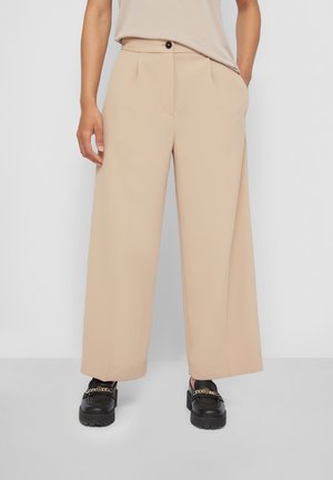 Persona con pantalones beige de pierna ancha y mocasines negros con detalle de cadena dorada, de pie frente a un fondo claro y liso.