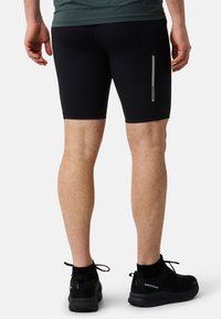 Shorts de sport noirs fabriqués dans un matériau extensible, avec une coupe ajustée, une bande réfléchissante sur le côté, portés avec des baskets noires.