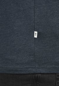 Dark navy shirt features a textured fabric with a side hem tag displaying an exclamation mark. The design is simple and casual.