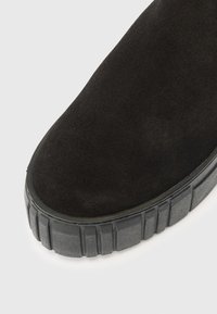 Klim na špičku černé semišové boty s tlustou vzorovanou gumovou podrážkou na světlém pozadí.