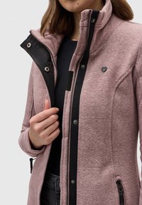 Veste rose clair avec un col haut, des accents noirs et une fermeture éclair visible. Comprend des boutons-pression, des poches latérales et un petit emblème sur la poitrine.