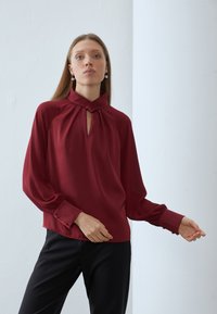 Burgundy silk blouse featuring a keyhole neckline, long sleeves with gathered cuffs, and a relaxed fit. Paired with black trousers.
