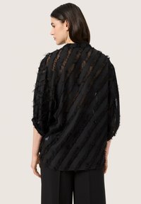 Top nero trasparente con righe verticali sfilacciate, vestibilità morbida, maniche a pipistrello e orlo curvo, sovrapposto a un tessuto leggero.