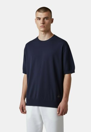 Giovane uomo che indossa un maglione corto oversize blu navy scuro e pantaloni bianchi, in piedi contro uno sfondo chiaro e uniforme.