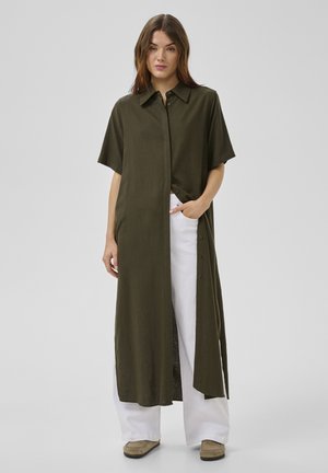 Nő hosszú, olívazöld, rövid ujjú, gombos ingruhával, fehér bő szárú nadrágban és bézs cipőben, egy kezét a zsebében tartva áll.