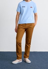 T-shirt azzurro chiaro a maniche corte con logo, pantaloni marroni sartoriali e scarpe slip-on a scacchi. Texture morbide e colori contrastanti visibili.