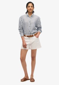 Camisa de botones a rayas azul y blanca, shorts blancos deshilachados, cinturón trenzado marrón y sandalias marrones de dedo. El modelo está de pie con las manos en los bolsillos.