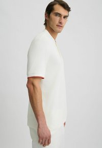 Cămașă cu mânecă scurtă de culoare crem deschis, cu manșete bordurate în roșu și o plăcuță galbenă. Textură netedă, croi lejer, design fără cusături.
