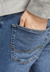 Denim jeans met een lichtblauwe wassing, met een achterzak met contrasterende gele stiksels en een hand die in de zak rust.