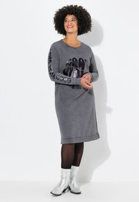 Robe grise à manches longues avec col rond, ornée d'un grand imprimé graphique et de texte sur les manches. Associée à des bottines argentées et des collants noirs.