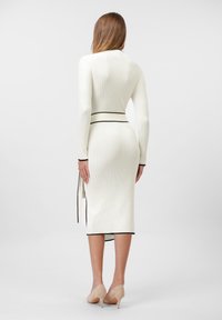 Knit dress in cream with a ribbed texture, featuring black trim along the neckline and hem, and a tied waist, paired with beige heels.