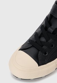 Basket en cuir noir avec lacets noirs, capuchon en caoutchouc crème, surface texturée et détails de couture contrastés.