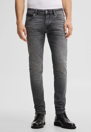 Mann trägt schmal geschnittene ausgewaschene graue Jeans und schwarze Leder-Dressstiefel und steht vor hellem Hintergrund.