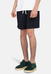 Marina bomullsshorts med dragsko i midjan, sidofickor och en avslappnad passform, kombinerat med gröna mockaskor med en beigefärgad sula.