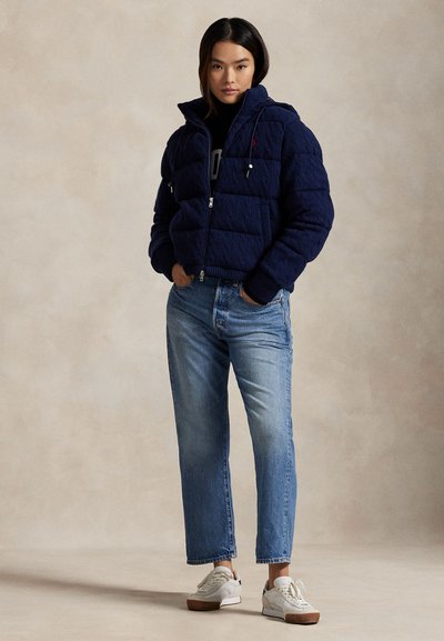 Mörkblå quiltad puffjacka med dragkedja och sidofickor, några ljusblå raka jeans och vita sneakers.