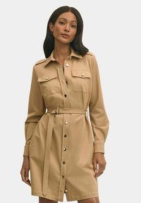 Abito camicia beige con maniche lunghe, due tasche sul petto, chiusura con bottoni, cintura in vita e texture liscia. Presenta dettagli con bottoni metallici.