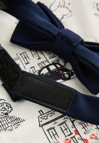 Nœud papillon bleu marine avec une bande Velcro noire reposant sur un tissu blanc imprimé de croquis noirs de maisons et de voitures et de fleurs colorées.