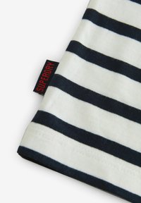 Triibuline valge ja tumesinine T-särgi kangas, millel on silt "Superdry" punases kirjas. Nähtavad õmblused äärtes. Pehme tekstuur.