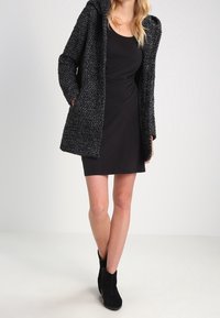 Svart stickad kappa med huva, texturerad design, buren över en figurnära svart klänning och svarta ankelskor, som framhäver en lagerlook.
