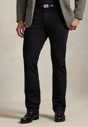 Uomo che indossa pantaloni neri, cintura nera, scarpe nere e una giacca grigia a spina di pesce con una camicia nera sotto.