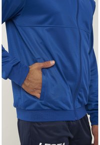 Giacca blu con zip, realizzata in tessuto testurizzato, dotata di tasca laterale e polsini a coste. Il design presenta una finitura liscia e una silhouette strutturata.