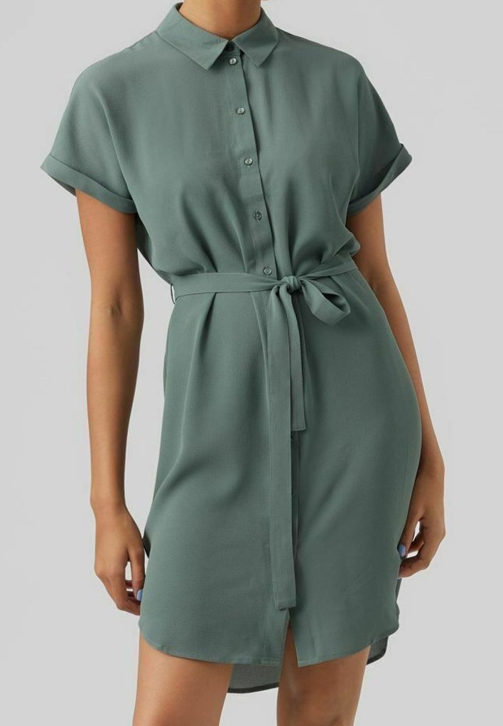 Robe chemise à manches courtes de couleur vert sauge, avec un col, une patte de boutonnage sur le devant, des manches retroussées et une ceinture nouée à la taille, portée par une personne debout.