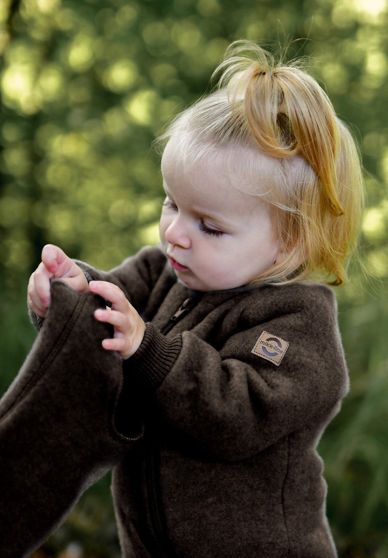Hnedá fleecová bunda so zipsom, rebrovanými manžetami a nášivkou s logom, ktorú nosí dieťa s blond vlasmi, ktoré si oblečenie prezerá.