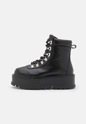Botte plateforme en cuir noir avec semelle épaisse à crampons, œillets métalliques, lacets noirs, col rembourré à la cheville et languette arrière, vue de côté extérieur.