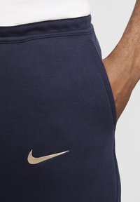 Navy joggerit pehmea tekstuuriga, mille vasakul küljel on kuldne Nike'i swooshi logo ja tihe pael.