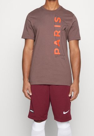 Homme portant un t-shirt marron avec le texte orange « PARIS » et un short Nike bordeaux avec le logo swoosh blanc ainsi que des chaussettes blanches montantes.
