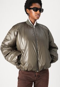 Olivgrüne gepolsterte Bomberjacke mit hohem Kragen, Reißverschluss und elastischen Bündchen. Glattes, glänzendes Material, kombiniert mit braunen Hosen.