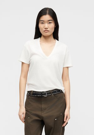 Fiatal nő fehér, V-nyakú, rövid ujjú pólóban, melyet barna nadrágba tűrt, fekete szegecselt övvel, egyszerű háttér előtt áll.