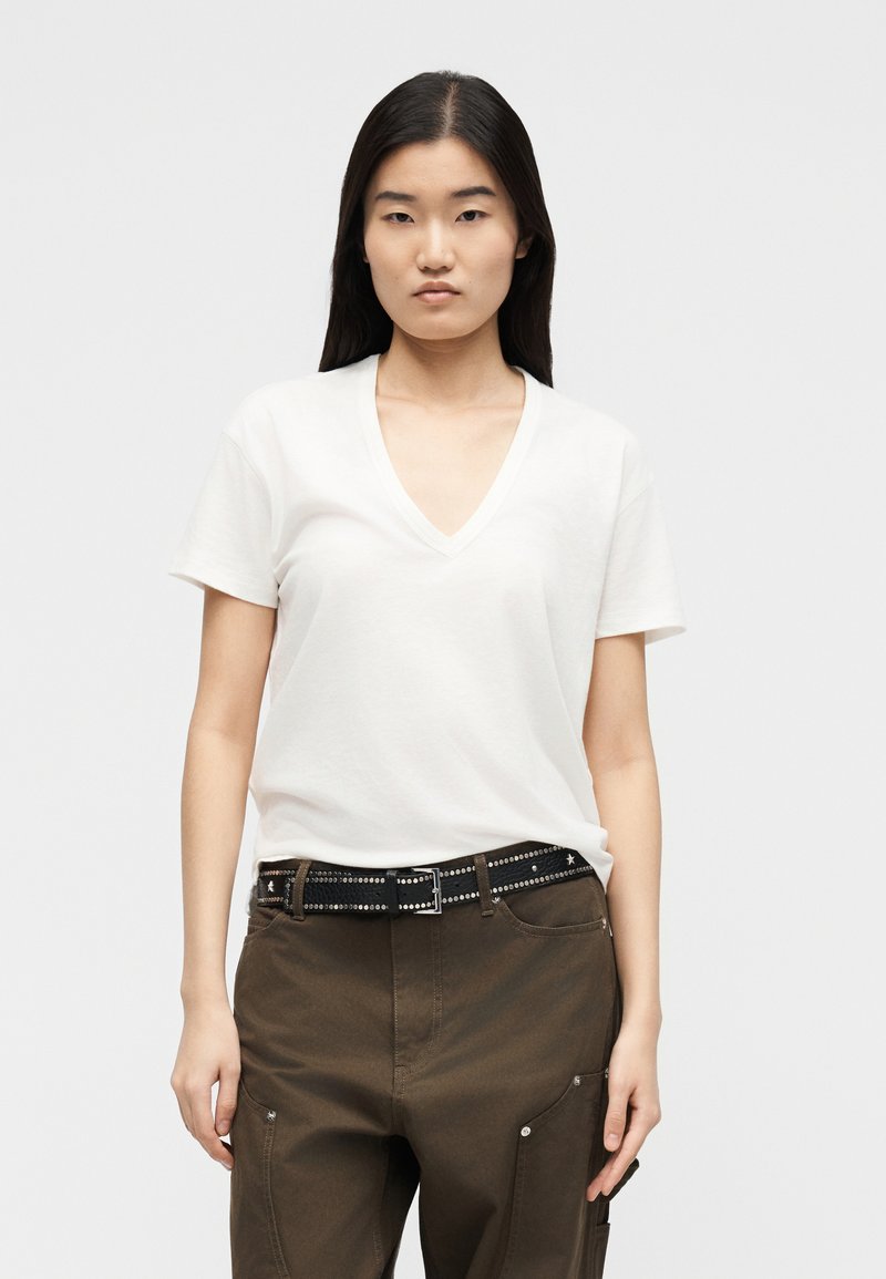 Νεαρή γυναίκα φορώντας ένα λευκό T-shirt με λαιμόκοψη V και κοντά μανίκια, μαζεμένο μέσα σε καφέ παντελόνια με μαύρη ζώνη με τρουκς, στέκεται μπροστά σε ένα απλό φόντο.