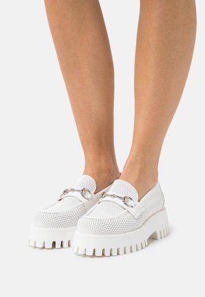 Mocassins en cuir blanc à plateforme épaisse avec une texture en mesh et un détail en mors de cheval argenté, portés sur des jambes nues devant un fond blanc.