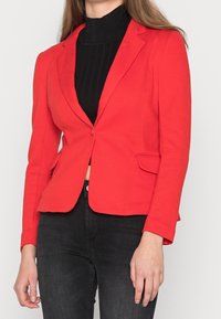 Femme portant un blazer rouge ajusté sur un pull à col roulé côtelé noir et un jean noir, posée avec les mains détendues le long du corps.
