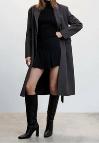 Donna che indossa un maglione nero a collo alto, una mini gonna plissettata nera, un lungo cappotto grigio e stivali neri con tacco alti fino al ginocchio, in piedi su un pavimento grigio.