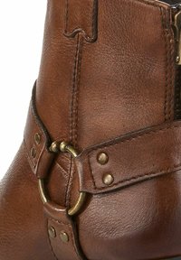 Bottine en cuir marron avec un bout arrondi, dotée d'une sangle décorative avec de quincailleries en laiton et des clous, une surface texturée et des détails cousus.