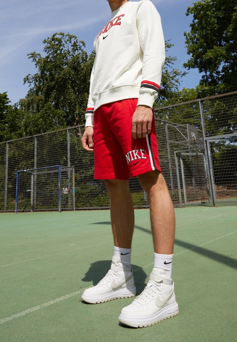 Osoba stojící na venkovním sportovním hřišti, oblečená v bílém svetru Nike, červených šortkách Nike, bílých vysokých teniskách Nike a bílých ponožkách Nike.