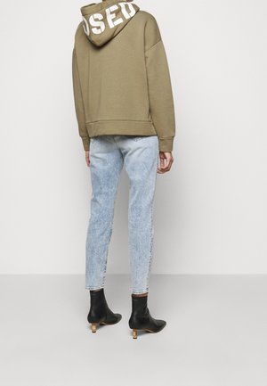 Personne portant un sweat à capuche beige avec un texte blanc sur la capuche, un jean bleu clair et des bottines noires à talons, debout devant un fond uni.