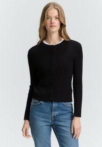Schwarze, gerippte Strickjacke mit langen Ärmeln, rundem Ausschnitt und frontaler Knopfverschluss. Kombiniert mit blauen Jeans, die ein tailliertes Design hervorheben.