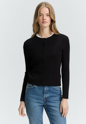 Schwarze, gerippte Strickjacke mit langen Ärmeln, rundem Ausschnitt und frontaler Knopfverschluss. Kombiniert mit blauen Jeans, die ein tailliertes Design hervorheben.