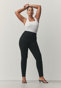 Mustad kõrge vöökohaga teksad, tiheda lõikega ja esitaskutega, mida kannab valge top ja mustad kontsaga sandaalid. Sile tekstuur.